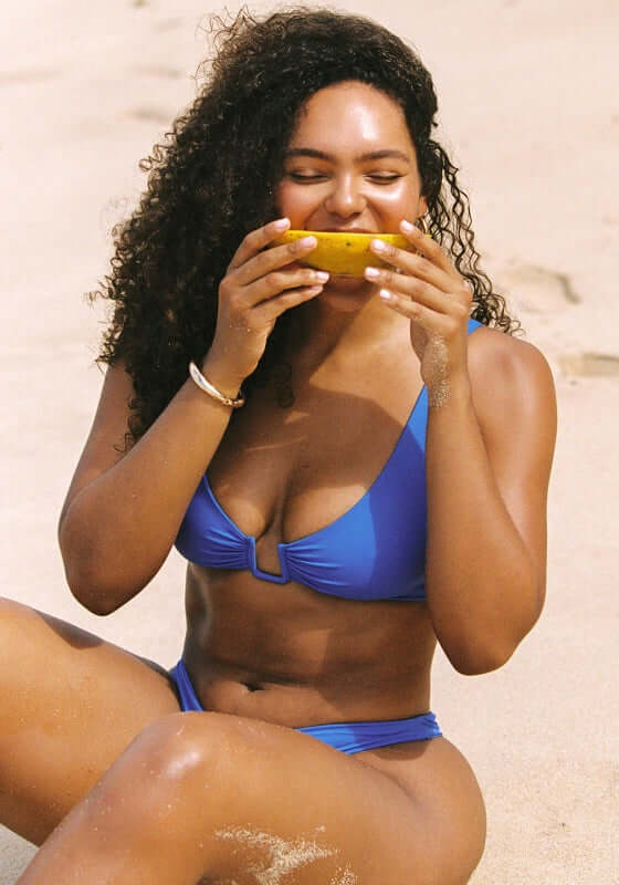 Palma Bikini Sicilian Blue by Away That Day model enjoying a snack on the beach, showcasing the chic bikini top and cheeky bottoms.