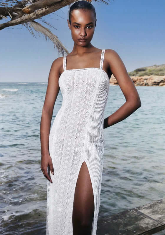 Vannie Long Dress White by Charo Ruiz, model wearing elegant dress with side slit by the beach.
