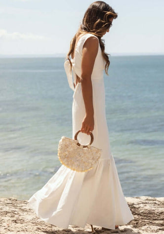 Fleur Dress White by Sole Lisboa, a model walks by the sea in a stylish white maxi dress with a bag.