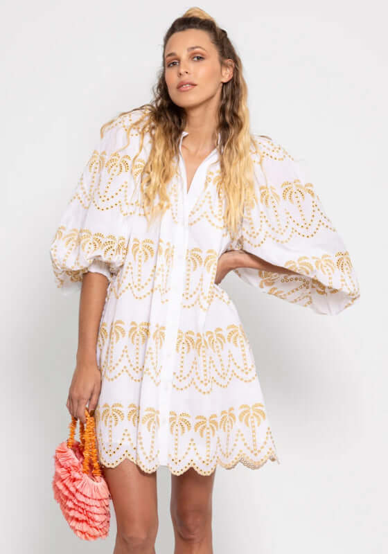 Inaya Dress by Sundress, model wearing a white palm tree eyelet sundress with puff sleeves and holding a pink handbag.