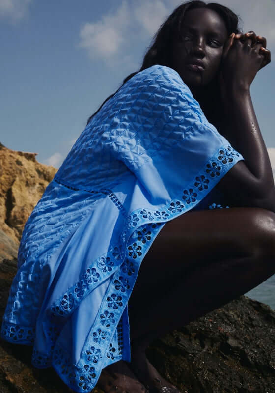 Maisie Kaftan Blue by Charo Ruiz, lightweight blue kaftan worn by a model sitting on a rock by the sea.