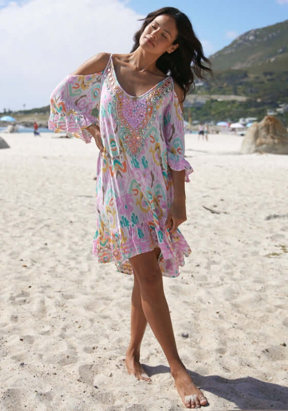 Birds Beach Dress by Miss June, a pastel pink dress with a paisley print and embellished neckline, worn on a sunny beach.