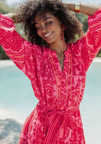 Samoa Dress Pink by Piti Cuiti features a woman smiling in a hot pink paisley dress with a belted waist and flowing sleeves.