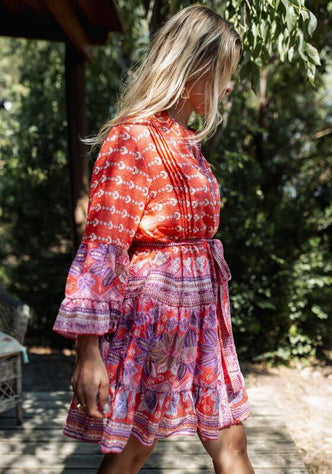 Curacao Dress Red Pink by Piti Cuiti, featuring a flowing skirt, tie belt, and peplum sleeves in vibrant red and pink hues.