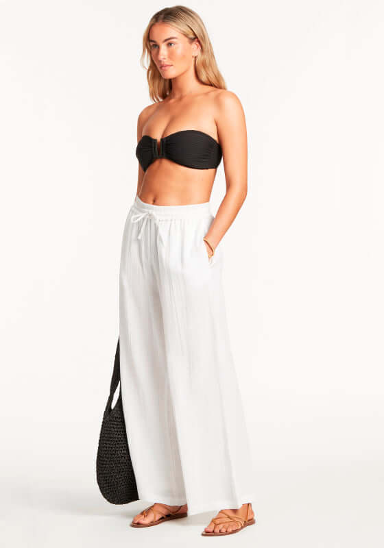 Sunset Beach Trousers White by Sea Level. Model wearing white wide leg beach trousers with black bikini.