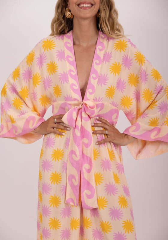Janis Dress Praia by Sundress, featuring a model in a vibrant floral print dress with kimono sleeves and a tied belt.