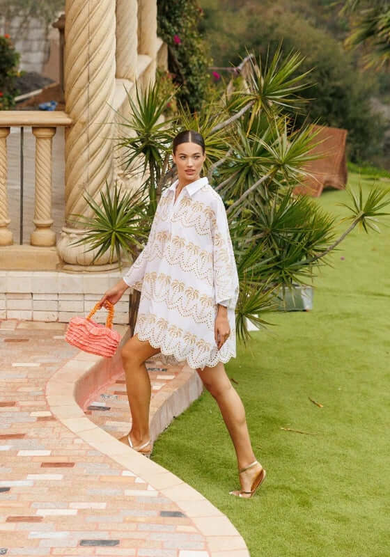 Flora Kaftan by Sundress, model wears a white shirt-like kaftan with gold details, holding a pink bag.