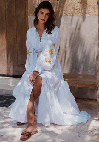 Nady Long Dress by Sundress, model in a white dress with embroidery, sitting on a bench in a tropical setting.