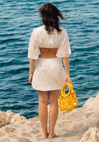 Severine Dress White by Sundress, showcasing a lightweight dress with cut out sides and a model holding a yellow bag by the ocean.