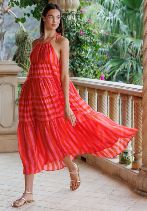 Valeria Dress by Sundress, model in bold pink and red striped long dress with flowing skirt on a balcony.