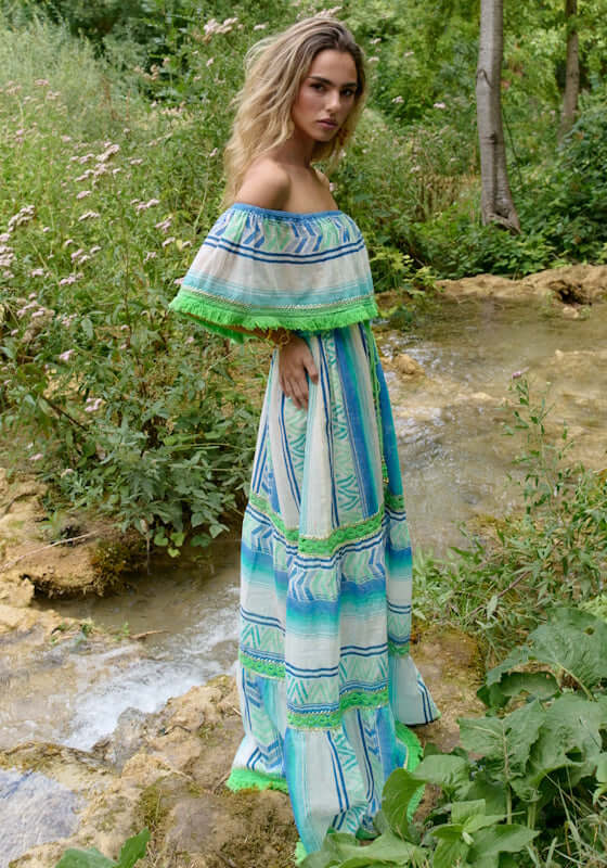 Frida Long Dress Aqua by Piti Cuiti, showcasing flowing silhouette, fringe trim, and vibrant aqua, blue, and green hues in a natural setting.