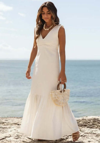 Fleur Dress White by Sole Lisboa, model in white maxi dress by the beach, featuring V-neckline and peplum skirt.