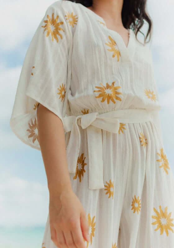 Dalhia Jumpsuit Coppacabana by Sundress, featuring shimmery gold sun motifs on a white jumpsuit with floaty sleeves.