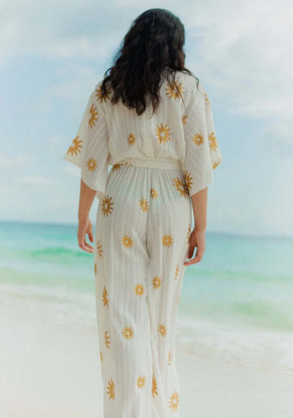 Dalhia Jumpsuit Coppacabana by Sundress, showcasing a model in a white jumpsuit with gold sun motifs against a beach backdrop.