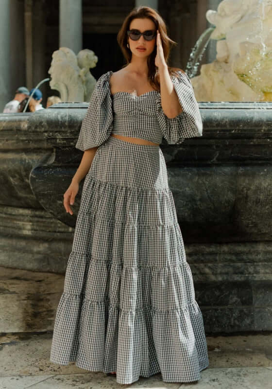 Odette Skirt Black Gingham by Sundress, a model wearing a tiered maxi skirt in black gingham at a fountain.