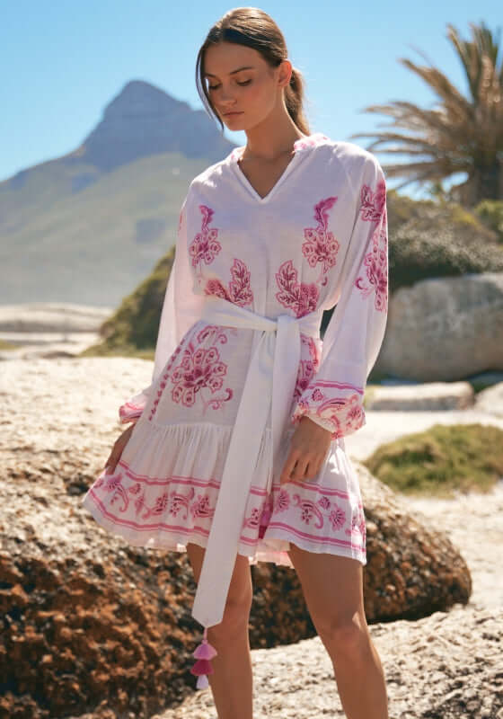 Anika Dress by Miss June, a charming pink paisley beach dress with V-neckline and tassel belt, set against a coastal backdrop.
