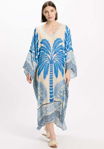 Rhoda Long Kaftan Blue by Miss June featuring a tropical palm print in Mediterranean blue on a beige background.
