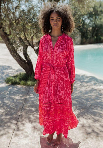 Samoa Dress Pink by Piti Cuiti, showcasing vibrant hot pink and red paisley print with flowing sleeves beside a pool.
