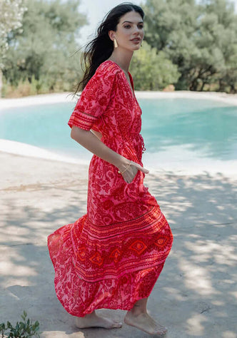 Samoa Short Sleeve Dress by Piti Cuiti worn outdoors, showcasing vibrant red and pink paisley fabric with a flowing maxi skirt.