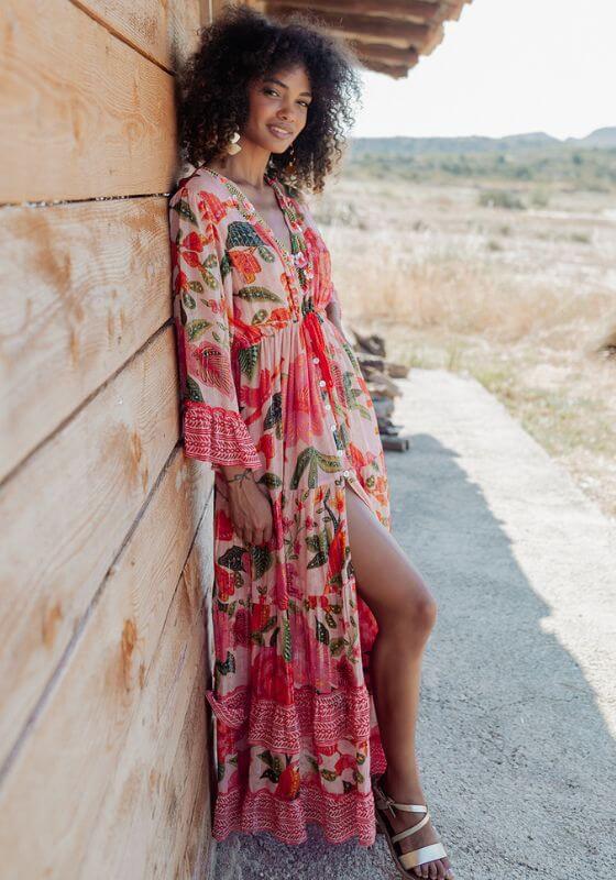 St Barths Beach Dress Orange by Piti Cuiti, vibrant orange dress with tropical prints, model standing beside wooden wall.