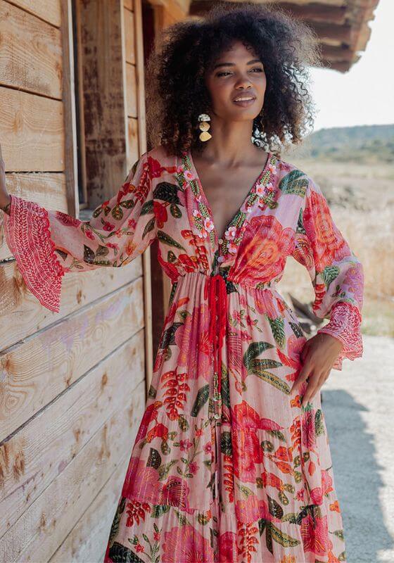 St Barths Beach Dress Orange by Piti Cuiti, a model showcases a vibrant floral dress against a wooden backdrop.