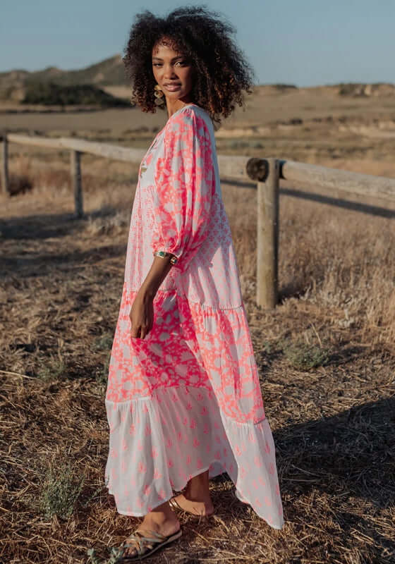 Ambar Long Beach Dress Orange Neon by Piti Cuiti worn outdoors, showcasing a vibrant pink and white tiered design.