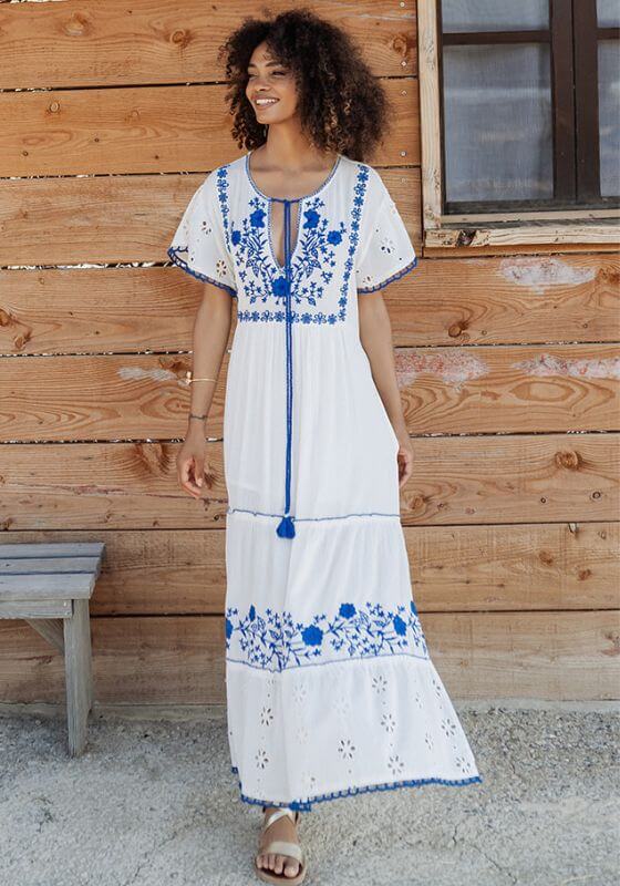 Tulum Long Dress Blue White by Piti Cuiti, showcasing a woman in a maxi dress with blue embroidery against a wooden backdrop.