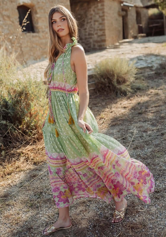 Curacao Dress Green by Piti Cuiti, a model wearing a flowy dress with ruffled collar and tassels in a garden setting.