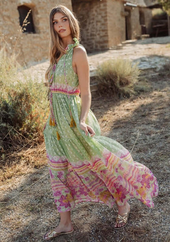 Curacao Dress Green by Piti Cuiti, a model wearing a flowy dress with ruffled collar and tassels in a garden setting.
