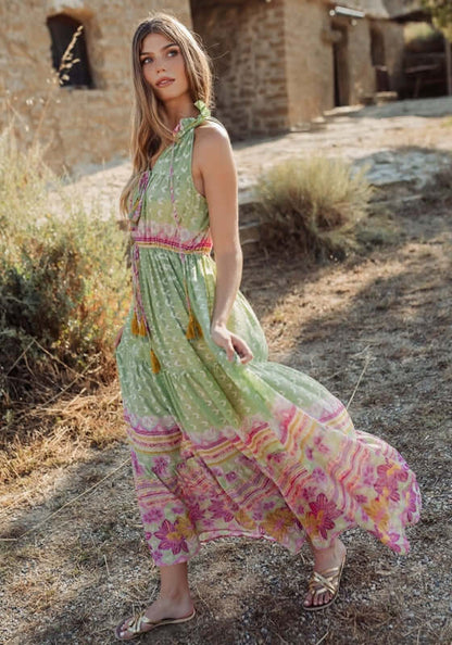 Curacao Dress Green by Piti Cuiti, a model wearing a flowy dress with ruffled collar and tassels in a garden setting.