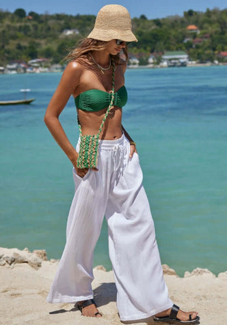 Sunset Beach Trousers White by Sea Level. Model in white wide-leg trousers, sunny beach, green bag, and hat.