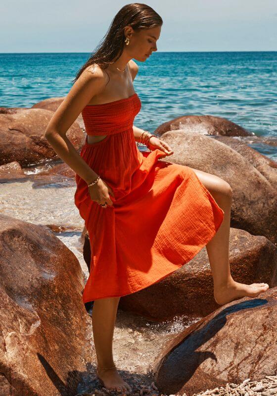 Sunset Bandeau Dress Flame by Sea Level worn by a model walking on the beach near rocks and water.