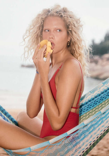 V Neck Swimsuit Cosmo Red by Seafolly, a model enjoying a snack in a hammock by the beach.